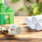 Thermostaatventiel en bouwtekeningen op een houten tafel met een groen huis van blokken, symbool voor duurzame verwarming met een warmtepomp.