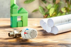 Thermostaatventiel en bouwtekeningen op een houten tafel met een groen huis van blokken, symbool voor duurzame verwarming met een warmtepomp.