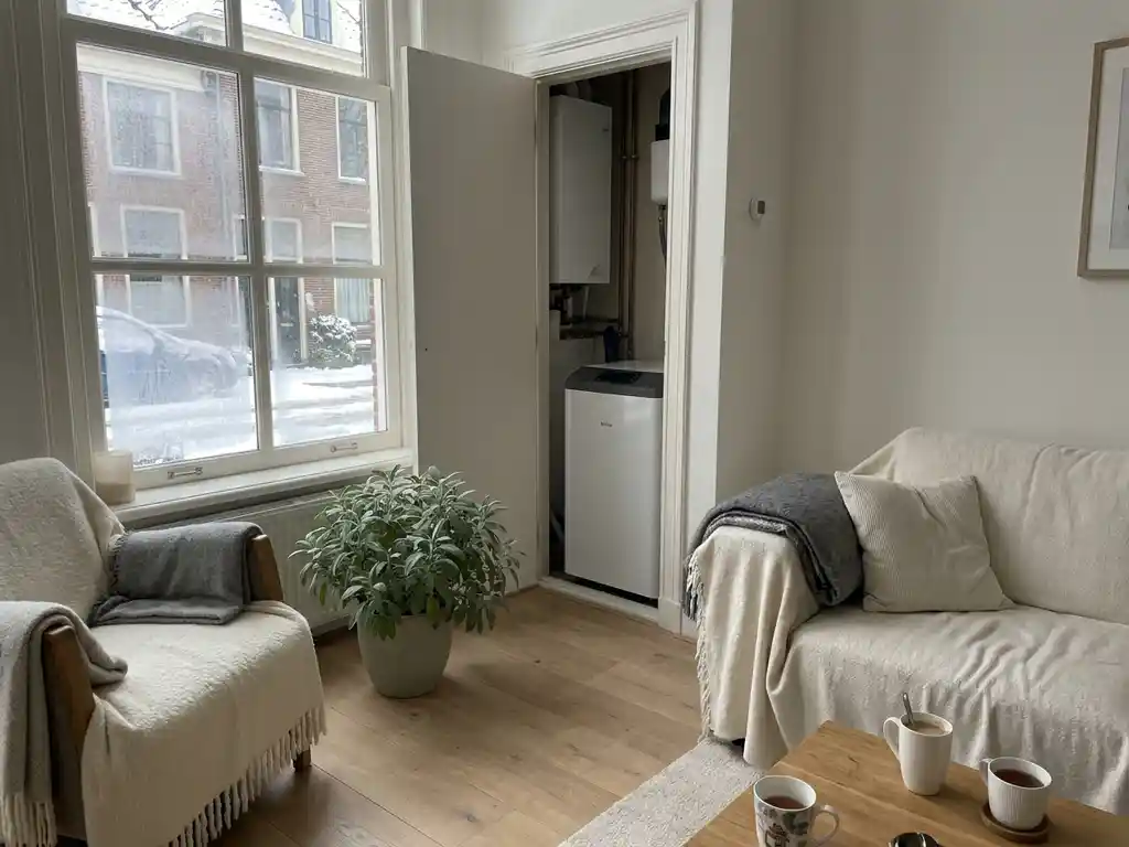 Gezellige Nederlandse woonkamer met lichte eikenhouten vloer en compacte witte warmtepomp in open kast, besneeuwd straatje zichtbaar door berijpt raam.